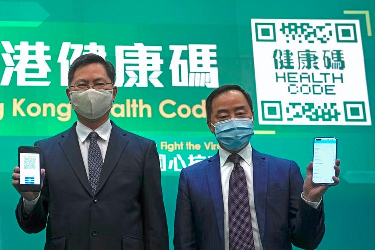 two Chinese officials hold up mobile phones as they stand in front of a green background with a QR code