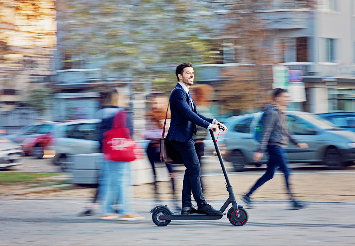 A man in a suit rides an e-scooter along a busy street in a city. He is smiling