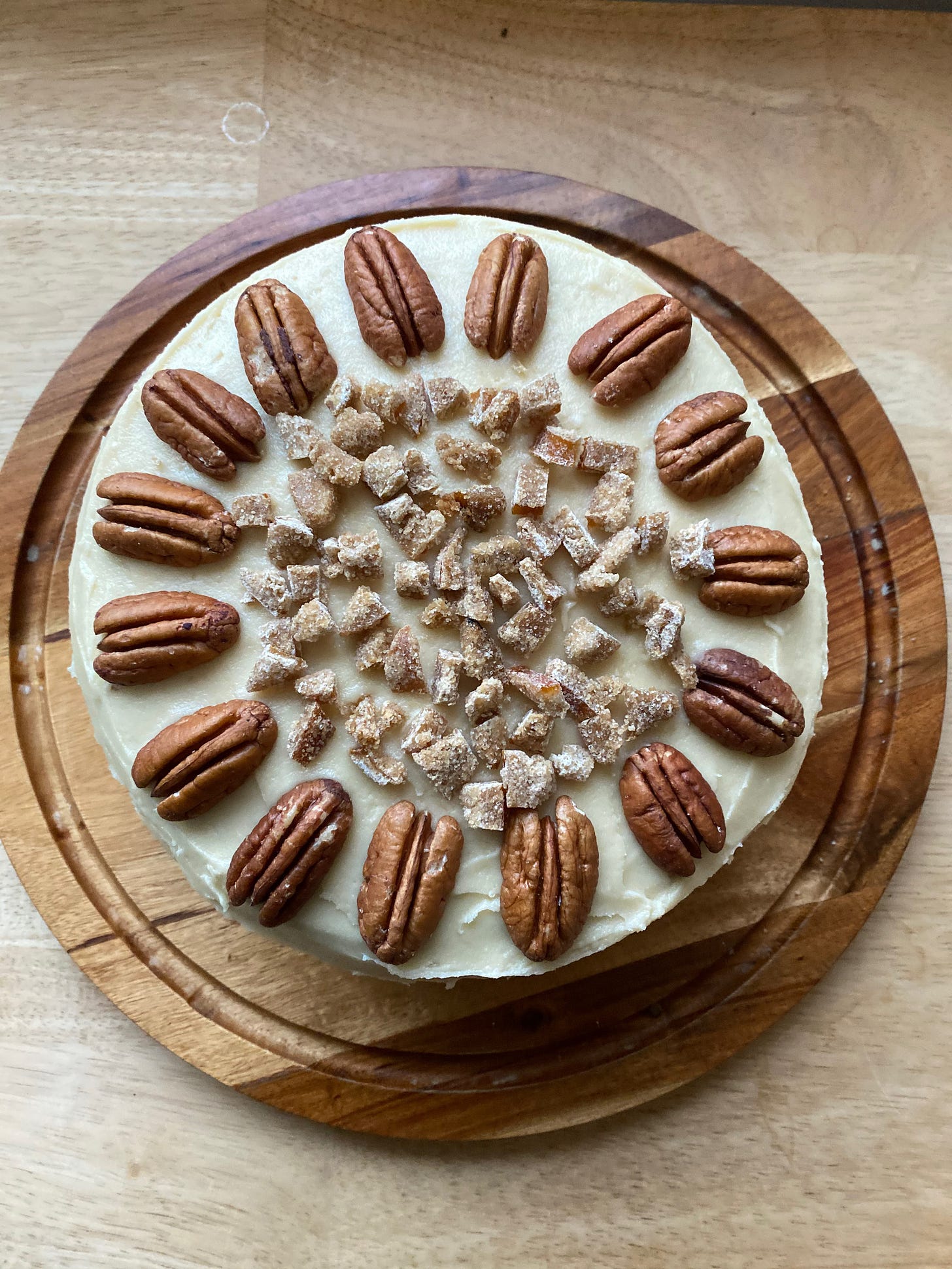 A round cake covered in creamy frosting sits on a wooden platter. It is decorated with a ring of pecan halves, and sprinkled with chopped candied orange peel.