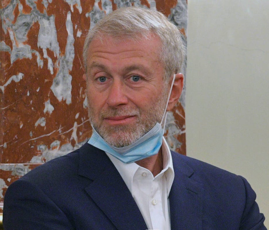 Photograph of Russian oligarch Roman Abramovich. He is looking slightly into the camera and smirking, wearing a blue covid mask hanging underneath his salt and paper bear. He is wearing a blue suit jacket and a white shirt with no tie.