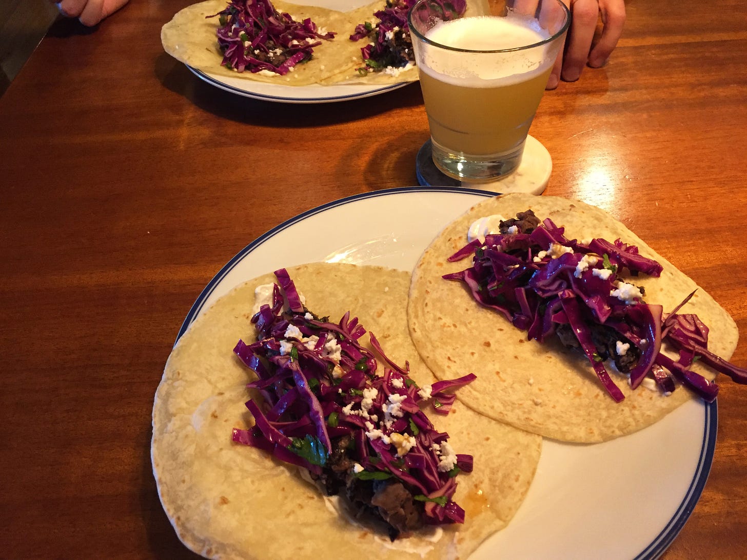 Two plates across from each other on a table, each with two toasted tortillas filled with black beans, feta, and red cabbage slaw. A glass of beer sits on a coaster between the plates.