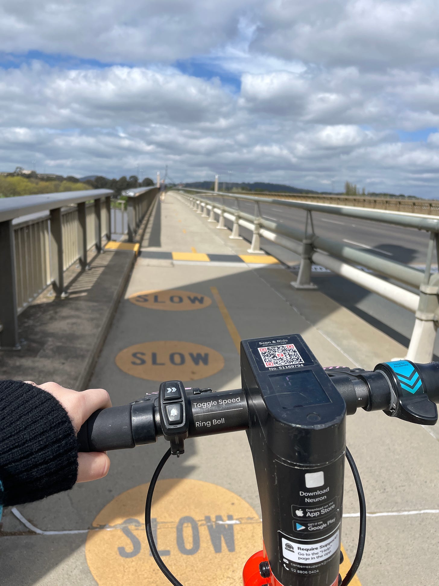 A view over the handlebars of an electric scooter. There are yellow circles with SLOW in black lettering on the path. The sky is cloudy and the spire of Australia's Parliament House is in the distance. 