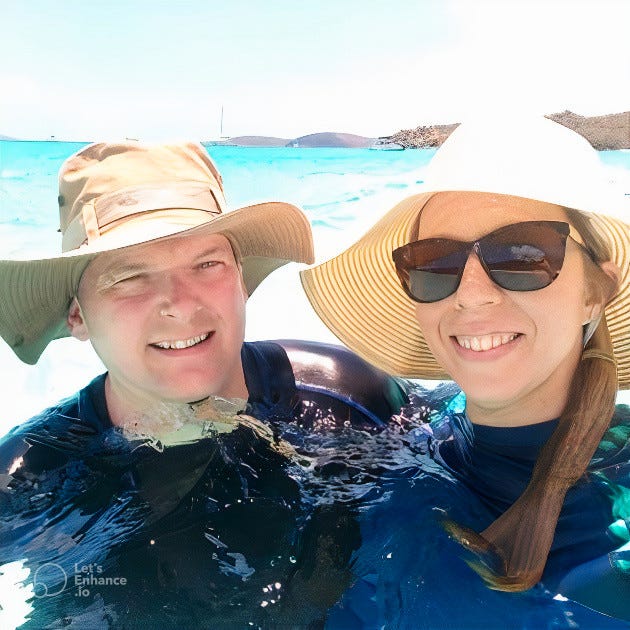 Bill and his wife are standing in the ocean wearing large beige sun hats and wet suits. Bill's wife is also wearing large sunglasses. Both are smiling into the camera. The sky in the background is pale blue.