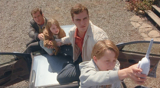 The Hess family as seen from above, with Morgan holding out a walkie talkie toward the camera/sky, Merrill behind him and Bo and Graham behind Merrill. They are all sitting on the hood or roof of the family station wagon.