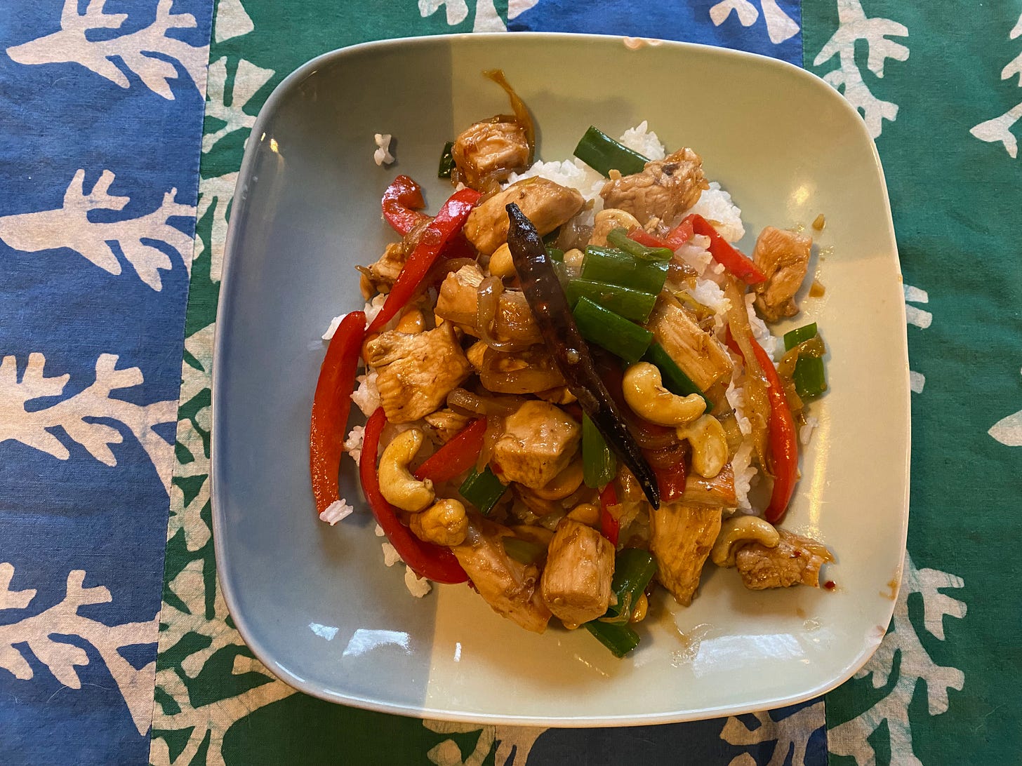A square plate full of stir-fry on a green and blue striped placemat. The stir-fry is full of chunks of chicken, cashews, strips of red pepper, and bright green scallions.