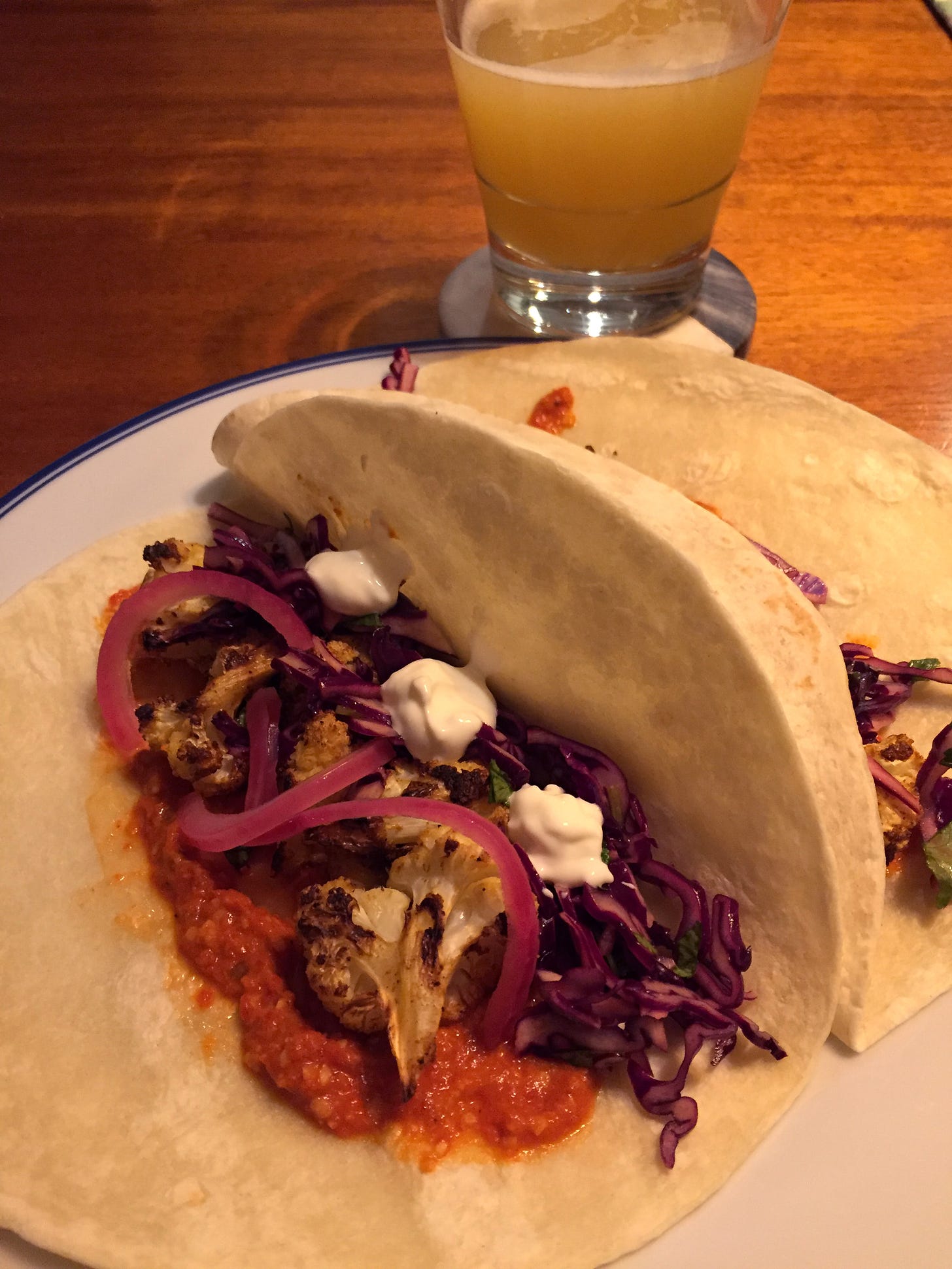 Two 8" flour tortillas filled with romesco, charred cauliflower, and red cabbage slaw. Dots of sour cream and pink slices of pickled onion sit on top, and a glass of hazy IPA rests on a coaster just to the upper right of the plate.