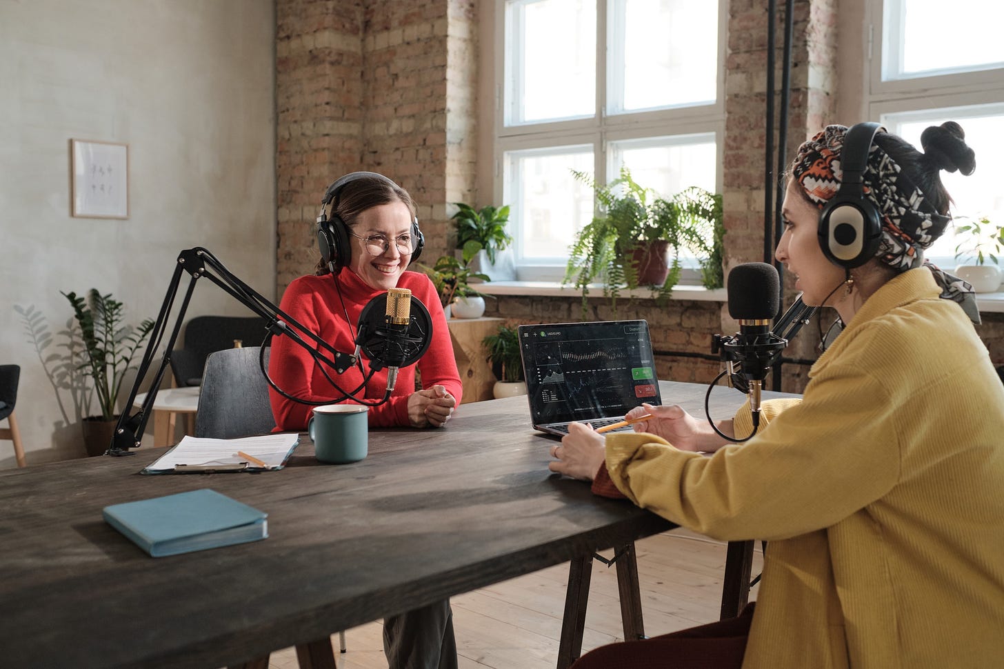 Two female podcasters casually speak to one another over microphones in a well-lit minimalist office .