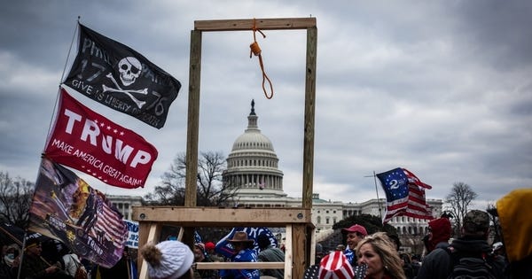 A photograph of the white supremacist insurrectionist riot that attempted to overthrow democracy in the U.S. The Capitol building is seen through the frame of a recently-constructed gallows.