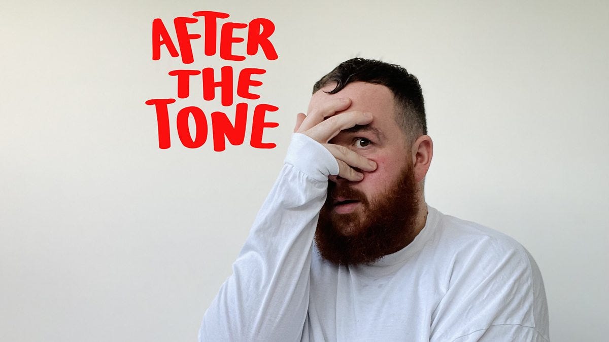 Performance artist Scottee is wearing a white long sleeve t-shirt, with short brown hear and a beard. He is holding his hand to his face but one eyes is peeping through his fingers. The backgroud is white. Above him, to the left are the words "After the Tone" in red letters