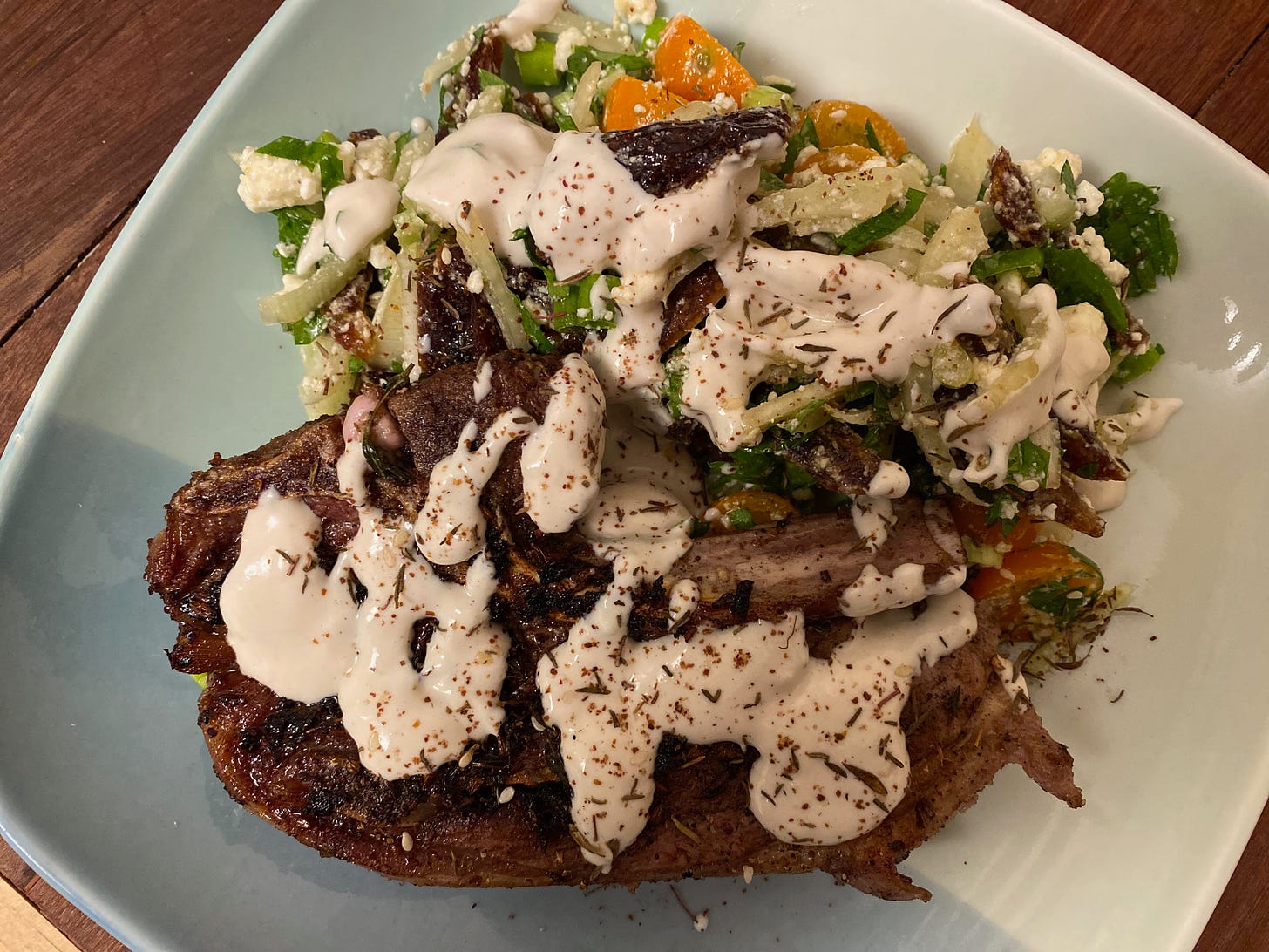 A square ceramic plate holding a browned lamb chop and a pile of fennel, date and cherry tomato salad. White tahini-lemon sauce is drizzled on top of the food, along with a sprinkling of ground sumac.