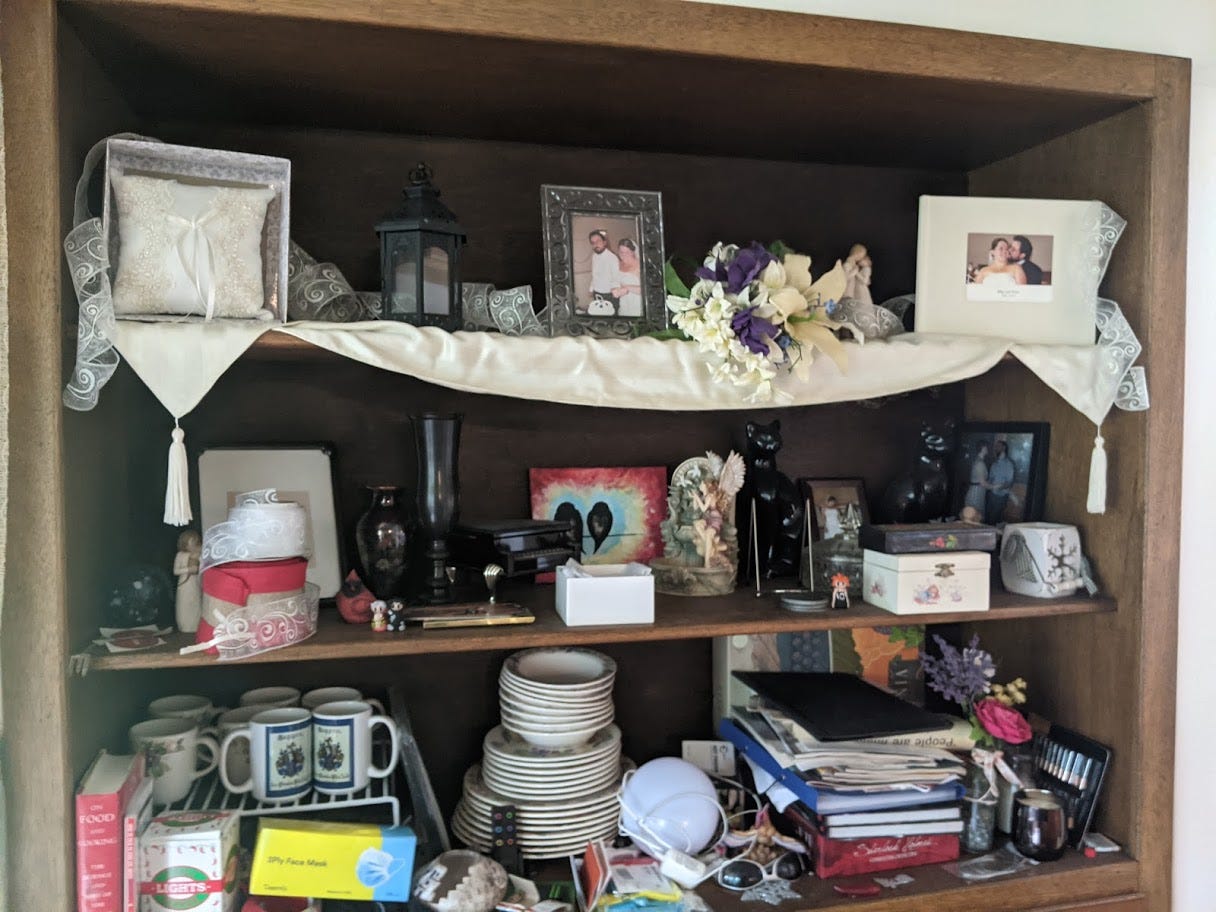 Several shelves, inlaid in a wall, with a variety of wedding-related keepsakes and photos, plus some art, some household curios, some nice dishes, a couple board games and books, and various piled junk, not quite chaotic, including a box of disposable masks