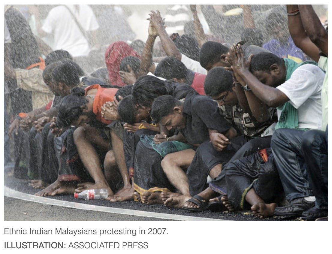 A picture of a group of dark skinned Indians crouched outside on the road in the pouring rain. Their heads are bowed and they are yelling something. Most of them are barefoot. Caption below picture reads: “Ethnic Indian Malaysian protesting in 2007.” Some of them have their hands raised with palms pressed together as if in prayer or Namaste.