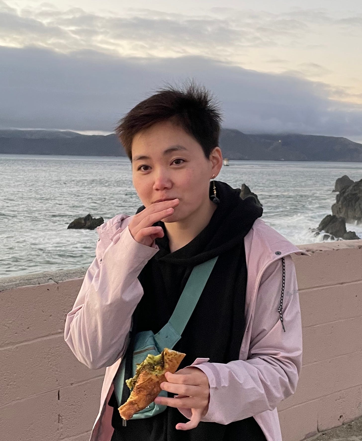 a person with short black hair standing outside and eating a slice of pizza. behind them is a seashore and cloudy sky.