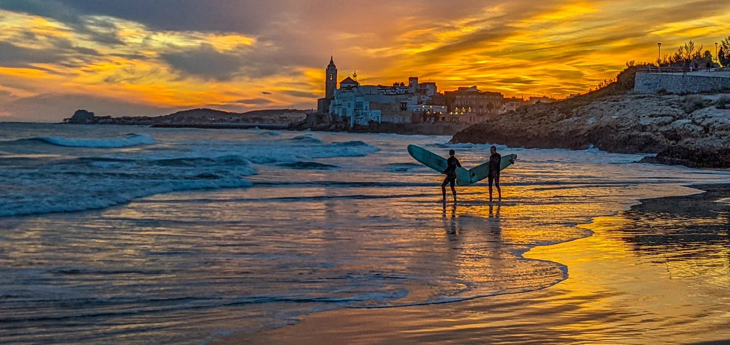 Two surfers in silhouette walking along the beach with both the water, sand, and sky golden from the sunset. 