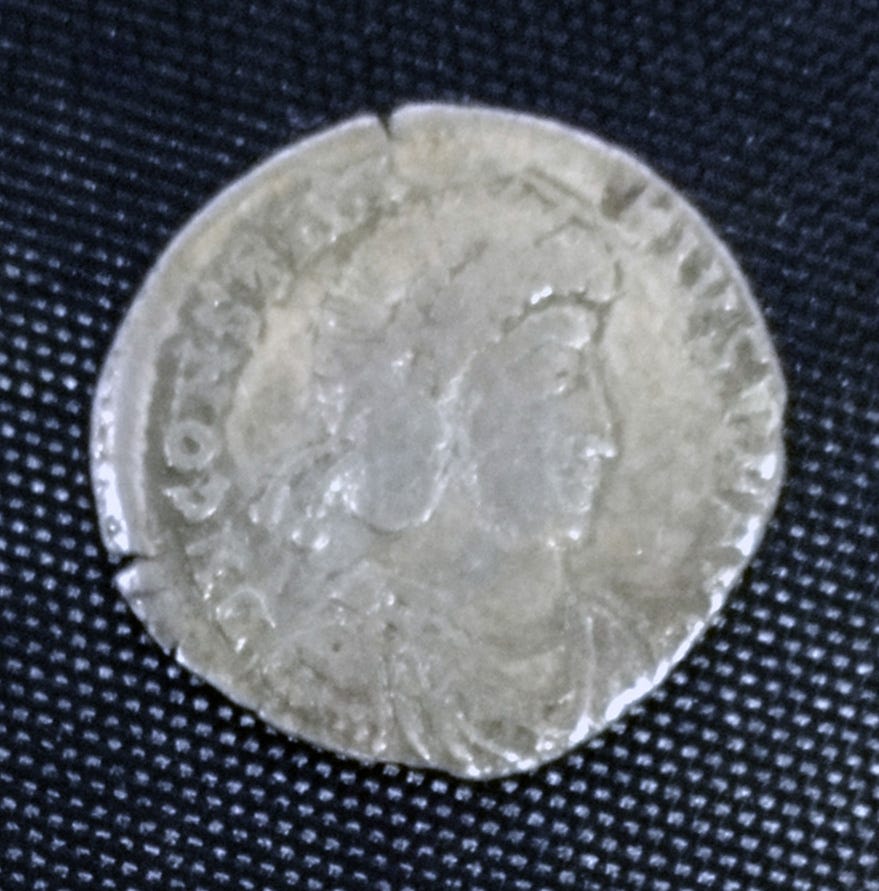 A silver coin in the centre a helmeted head surrounded by a circle of inscription with edging between that and the rim.