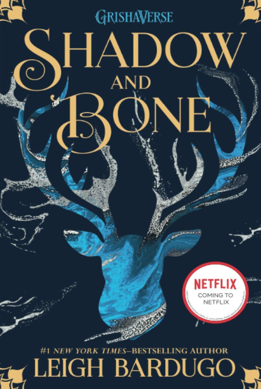 The navy blue cover of Shadow and Bone by Leigh Bardugo, depicting a light blue and gray stag and the rest of the text in gold, swirling font. 