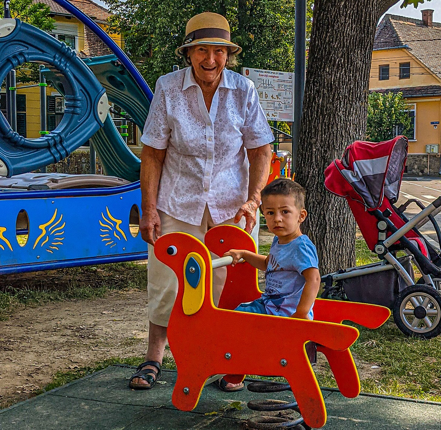 A smiling grandmother stands with her three-year-old grandson sitting on a rocking chair.