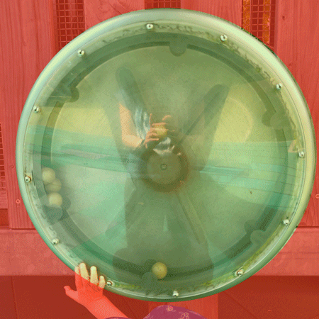 Rainbow Squared Year 5, Piece Twenty-Seven, 4. Red Green. An animated loop of a transparent plastic spinning wheel with three small balls flying around inside of it. It is on a playground structure, and in the photo you can see the reflection of the hands holding the camera and people walking by. As the loop restarts, a small hand reaches up to spin the wheel again. E’s small hand. The background surrounding the circle has been overlaid with red and the center has been overlaid with green. 