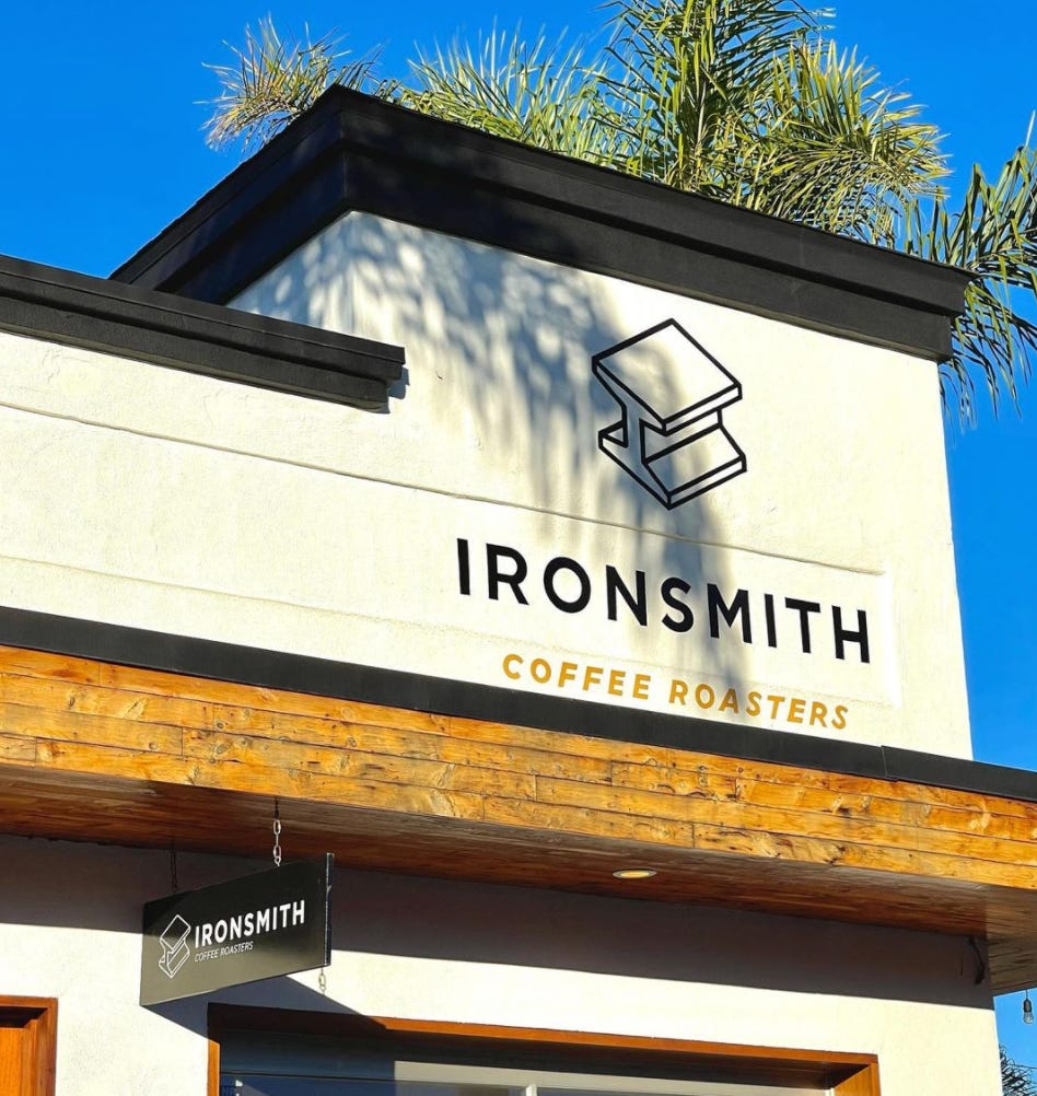 A front profile of Ironsmith Coffee Roasters entrance. A white building with black trim and the Ironsmith Coffee logo above the door. At the top a palm tree framed by blue sky pokes over the roof line.