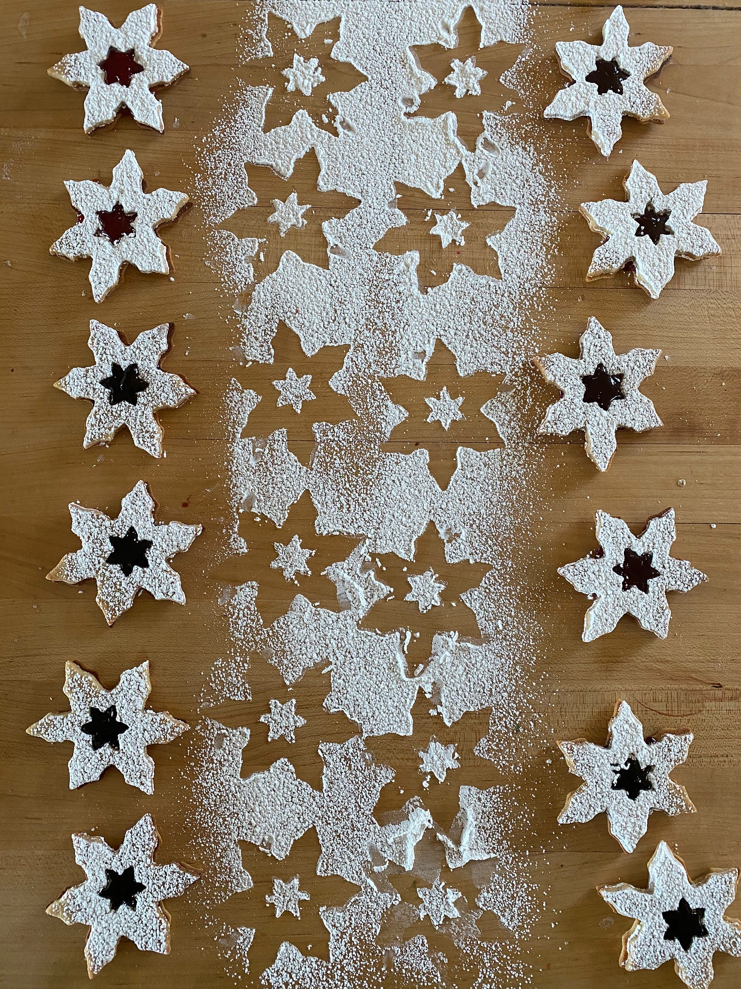 Two rows of star-shaped Linzer cookies on a wooden counter. They are coated in powder sugar, and each one has a small star-shaped opening through which the jam filling is visible. Between the two rows of cookies, the counter is covered in powdered sugar, showing the star-shaped outlines of where the cookies were.