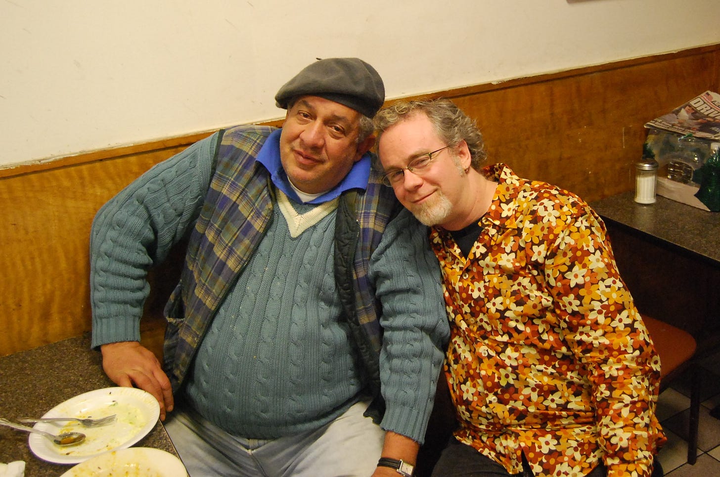 A white man with a chin beard wearing a flowered shirt rests his head on the sholder of a larger Egyptian man wearing a beret, a plaid vest, and a V-neck sweater. Empty plates at a table in front of them indicate they are in a restaurant.