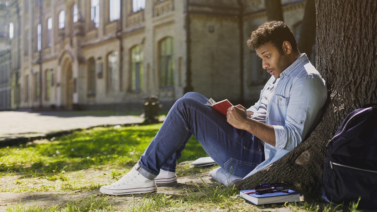 Multinational young male sitting under tree, reading gripping book, surprised