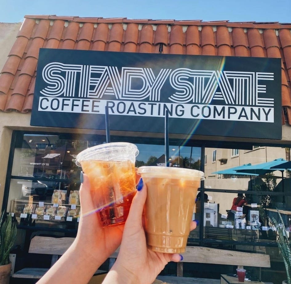 Two clear plastic cups with straws containing an iced tea and a cold brew coffee being held by women's hands held up in front of the entrance to Steady State Coffee Roasting. The Steady Sign (white on black) is the primary background.
