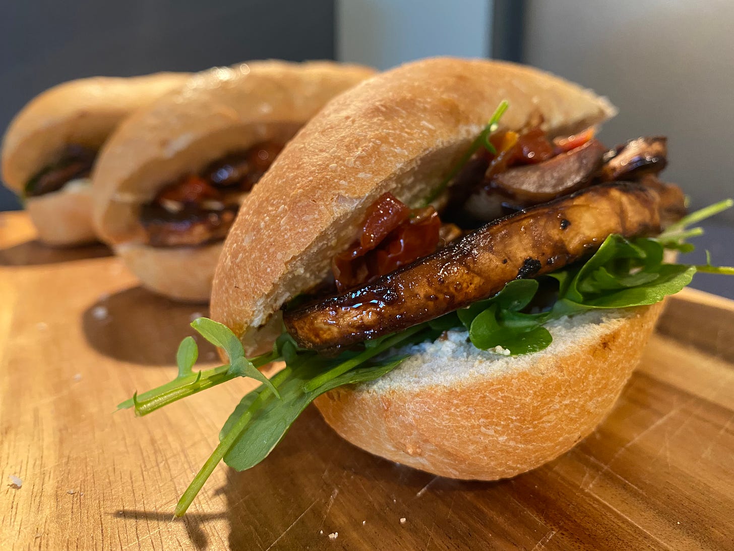 Row of three baguette sandwiches on a wooden chopping board. Each roll is filled with mushrooms, salad leaves and sun-dried tomatoes.