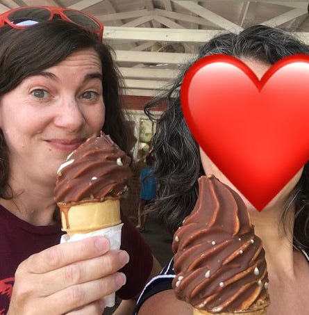 Emily, a white woman with dark hair, holds a Dole Whip dipped in chocolate. Her friend Jamie's face is covered with a big red heart