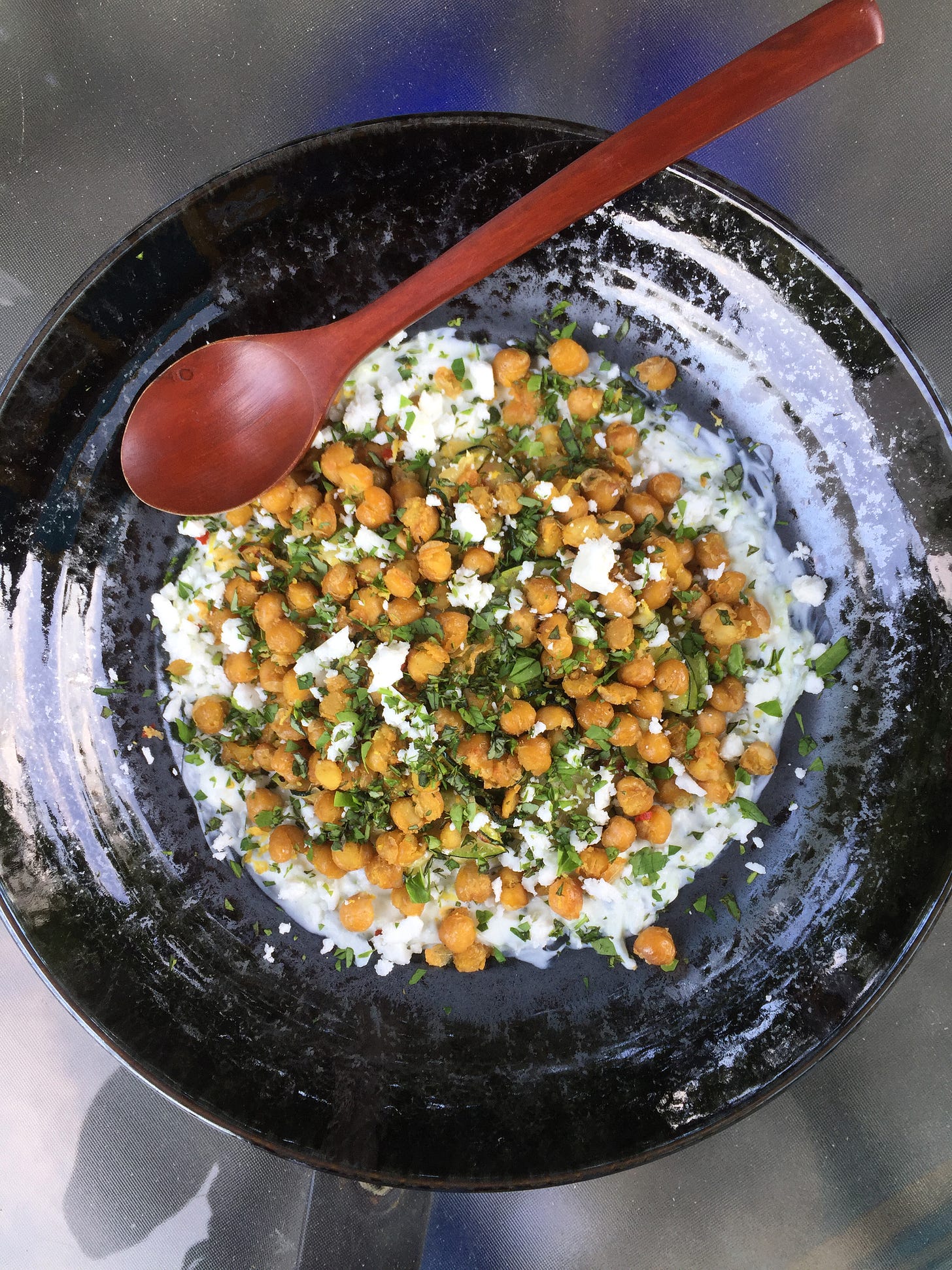 from above, a shallow black plate filled with tzatziki topped with zucchini, fried chickpeas, and a scattering of herbs and crumbled feta. A wooden spoon sits an an angle near the top.