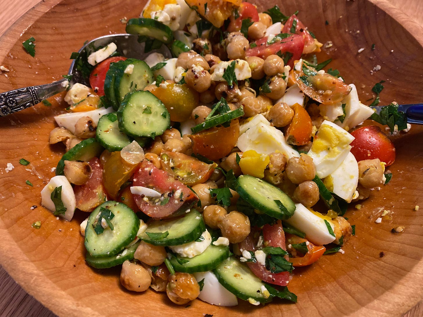 A wooden bowl filled with salad of chickpeas, cucumbers, bright red and orange cherry tomatoes, and hardboiled eggs.