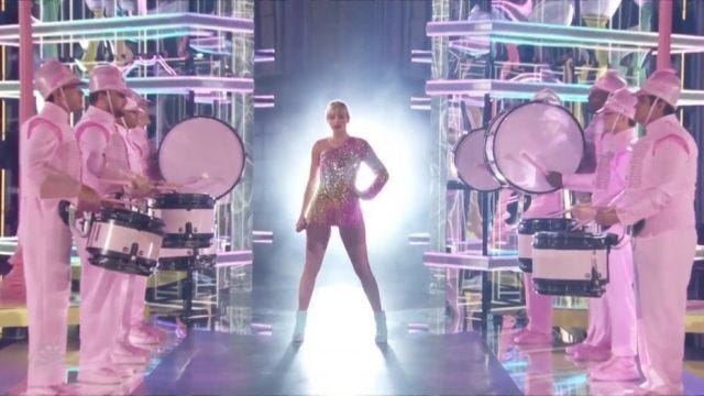 Taylor Swift on a stage in a pink sparkly leotard, surrounded by mixed gender drummers in pink uniforms. Taylor Swift on a stage in a pink sparkly leotard, surrounded by mixed gender drummers in pink uniforms.