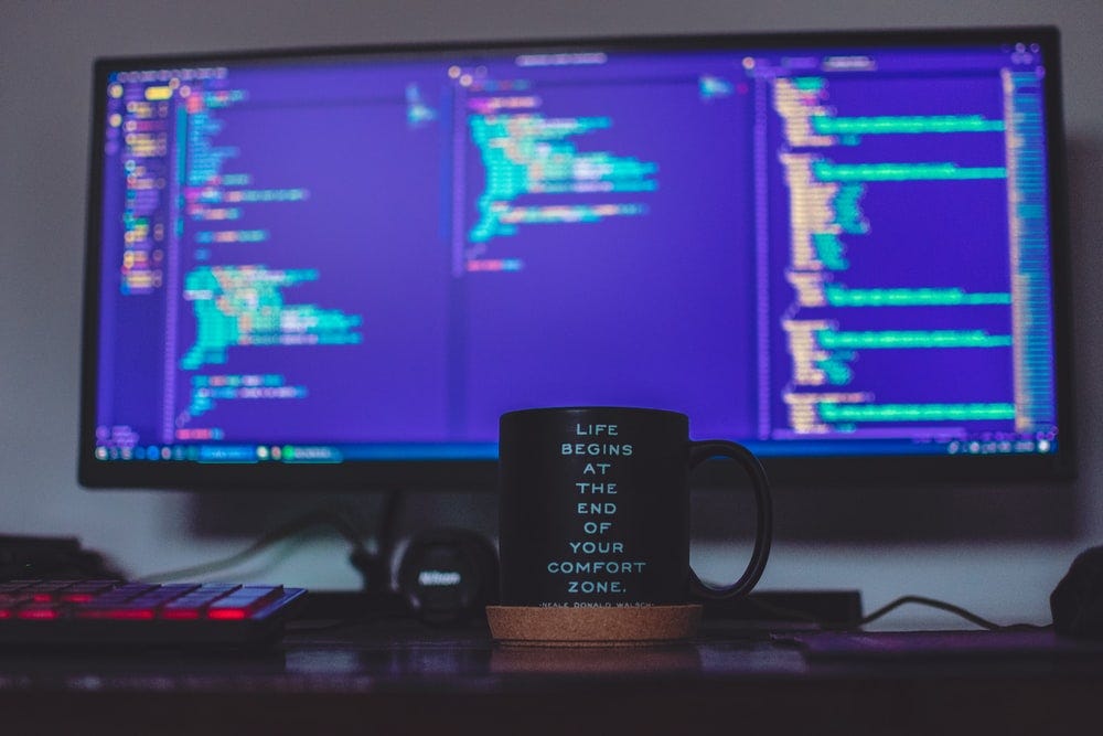 A coffee mug sits in front of a monitor showing a code editor. The mug has written on it: "Life begins at the end of your comfort zone."