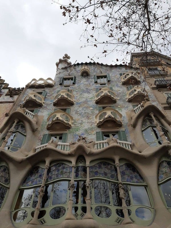 Fachada da Casa Batlló, construção de Antoni Gaudí em Barcelona. Foto de Edmara Galvão. Descrição da Imagem: é um edifício de cinco andares, repleto de curvas e sacadas e azulejos azuis coloridos. No primeiro andar, uma grande varanda repleta de vidros e vitrais azuis fica de frente para a rua, um andar acima. Nos andares superiores, de cada uma das quatro janelas que surgem, a varanda tem um formato branco misterioso — podem remeter a máscaras, ou caveiras. No último andar, um balcão branco e individual solitário desponta.