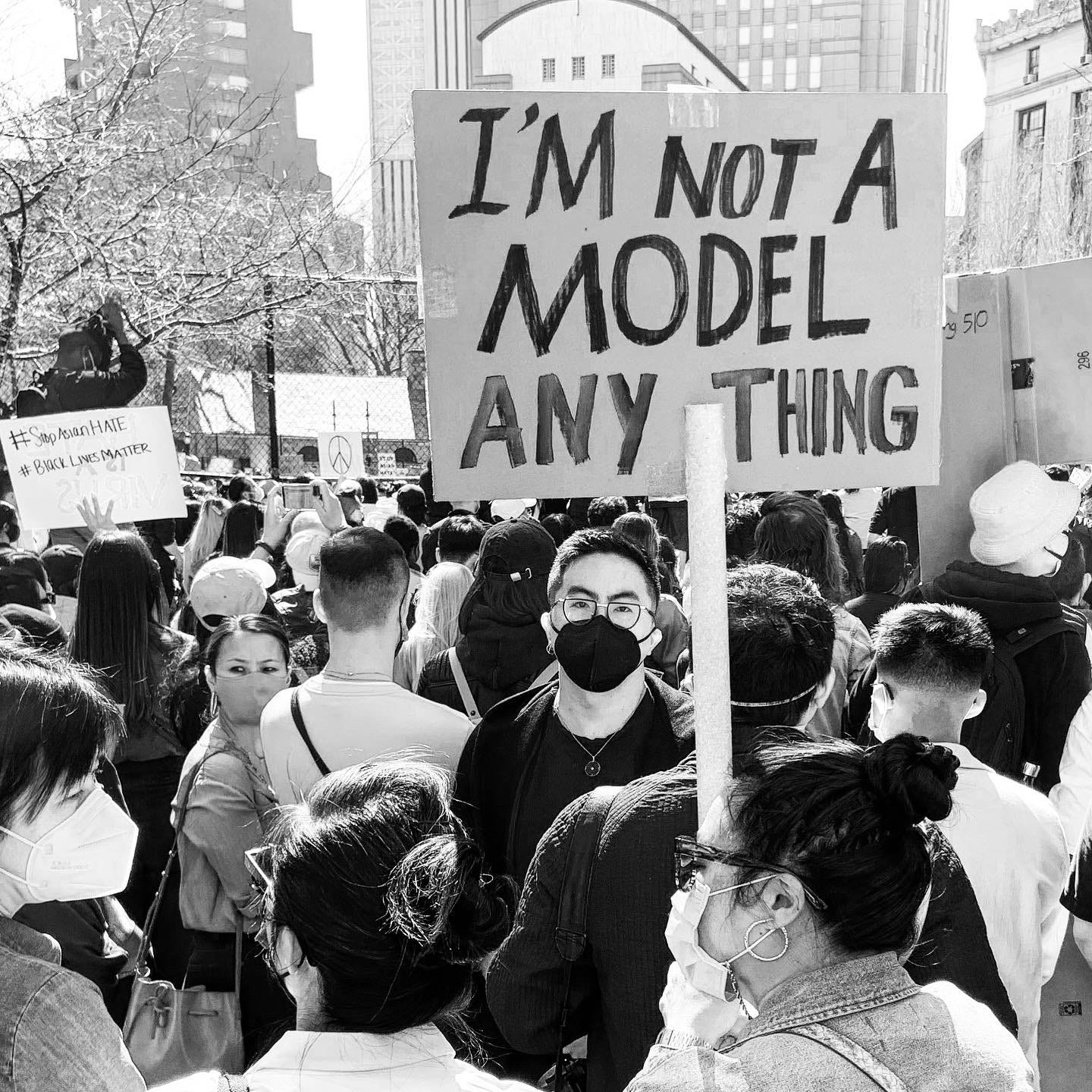 photo of a crowd at a rally to stop Asian hate; person in foreground holds a sign that reads "I'm not a model anything"; Bowen Yang stands directly underneath the sign, staring at the camera