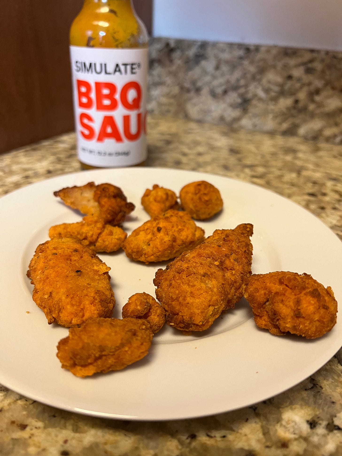 A plate of various nugget-like chicken wings, with a bottle of BBQ sauce behind them.