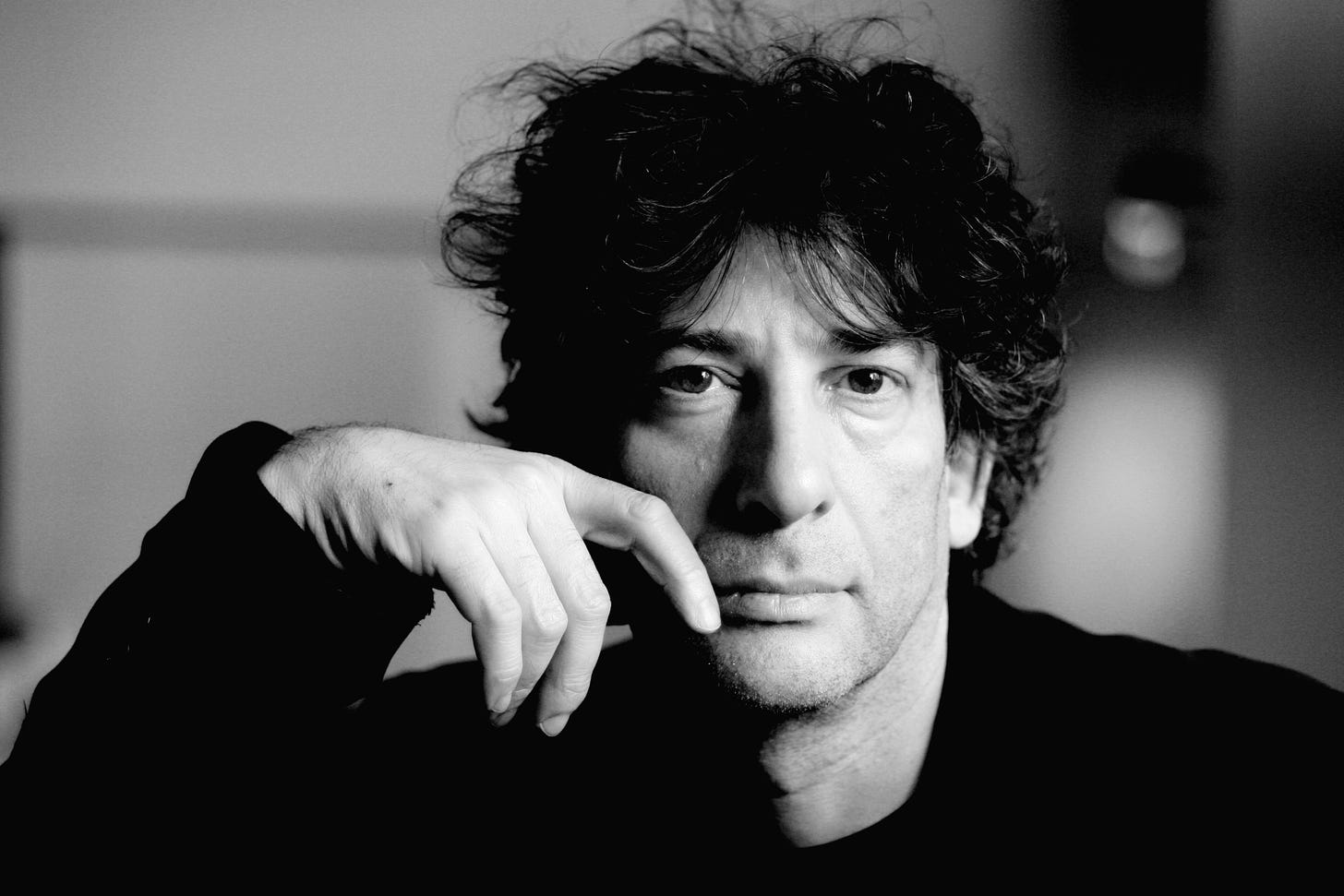 Foto preto e branca de Neil Gaiman olhando para frente, a mão apoiada na frente do rosto. Ele usa preto, tem o cabelo preto despenteado, com alguns fios brancos aparecendo.
