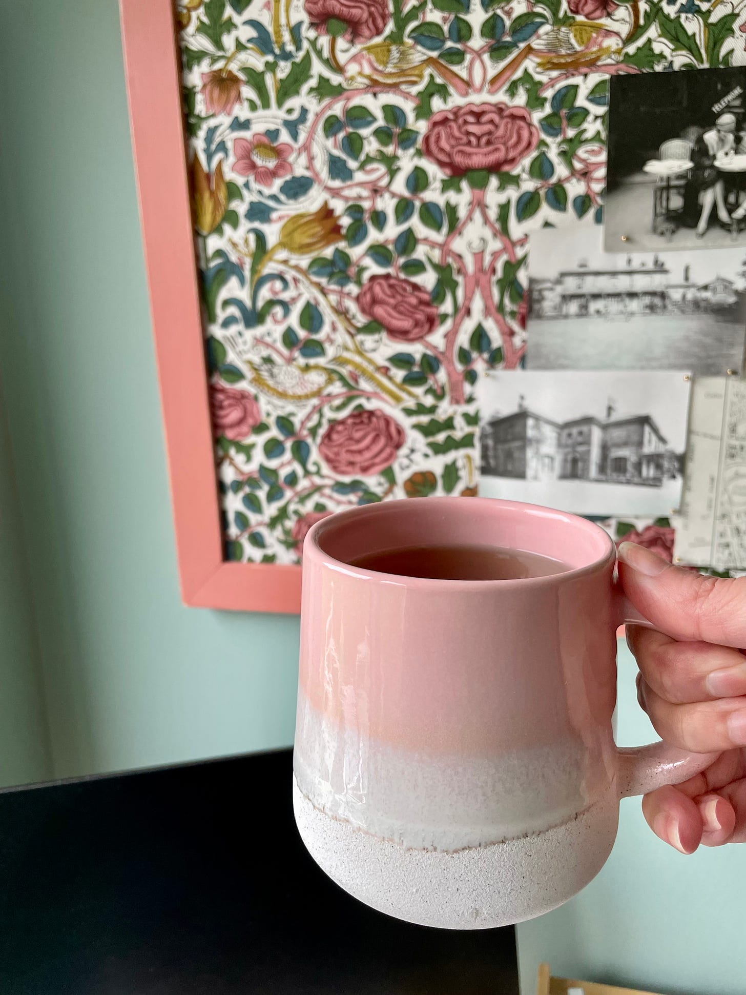 A pink mug held above my desk. Behind it you can see a monitor screen, the green walls and a floral pinboard. The pinboard has some black and white inspiration photographs on it, mainly of country houses.