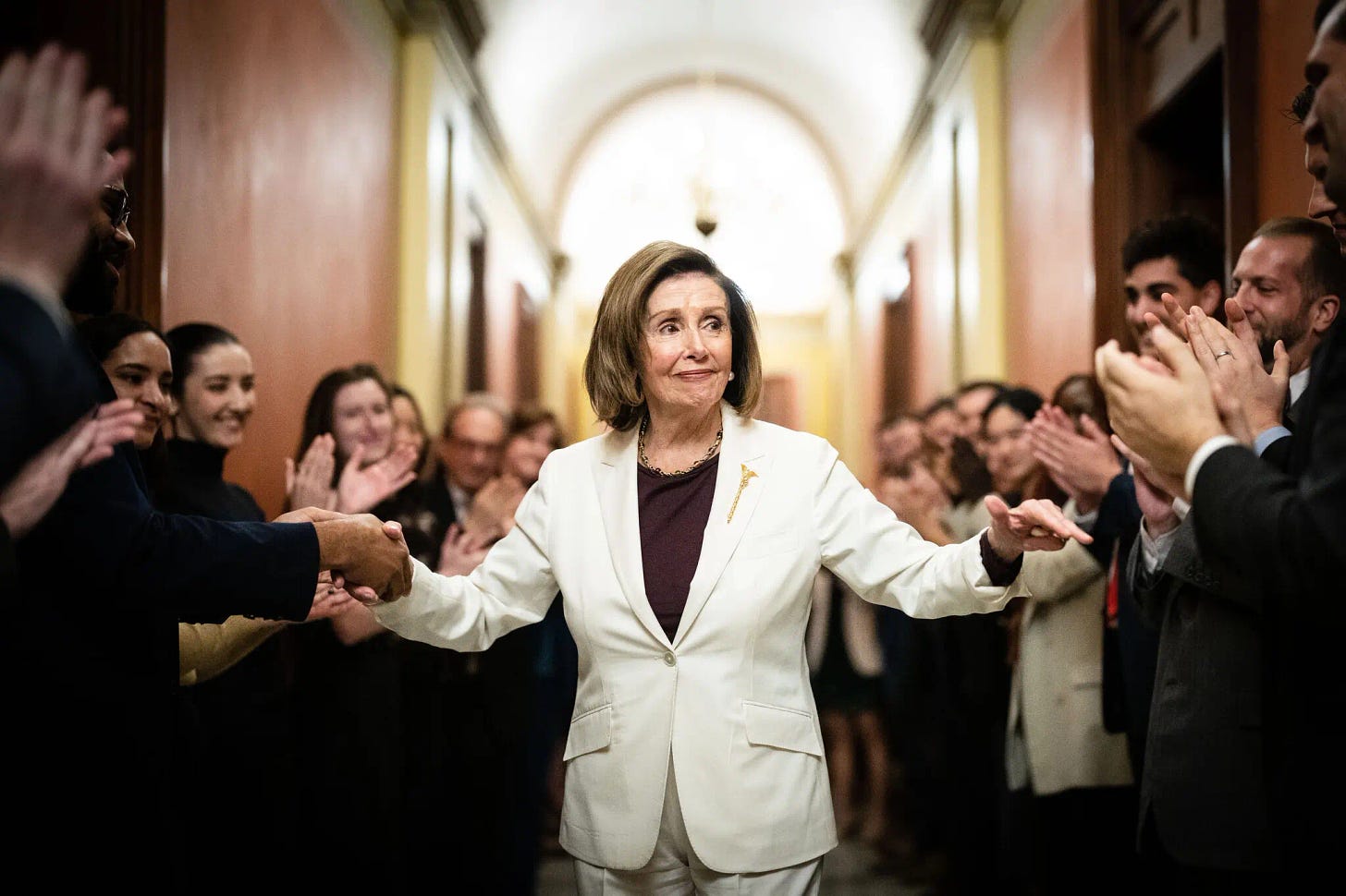 Nancy Pelosi, in a white pant suit, walks down a long hallway in the US Capitol, both of her arms outstretched to accept the handshakes and applause of the people lining the hallway. The hallway is filled with them, and the clap and smile as Pelosi walks, a wry smile on her face.