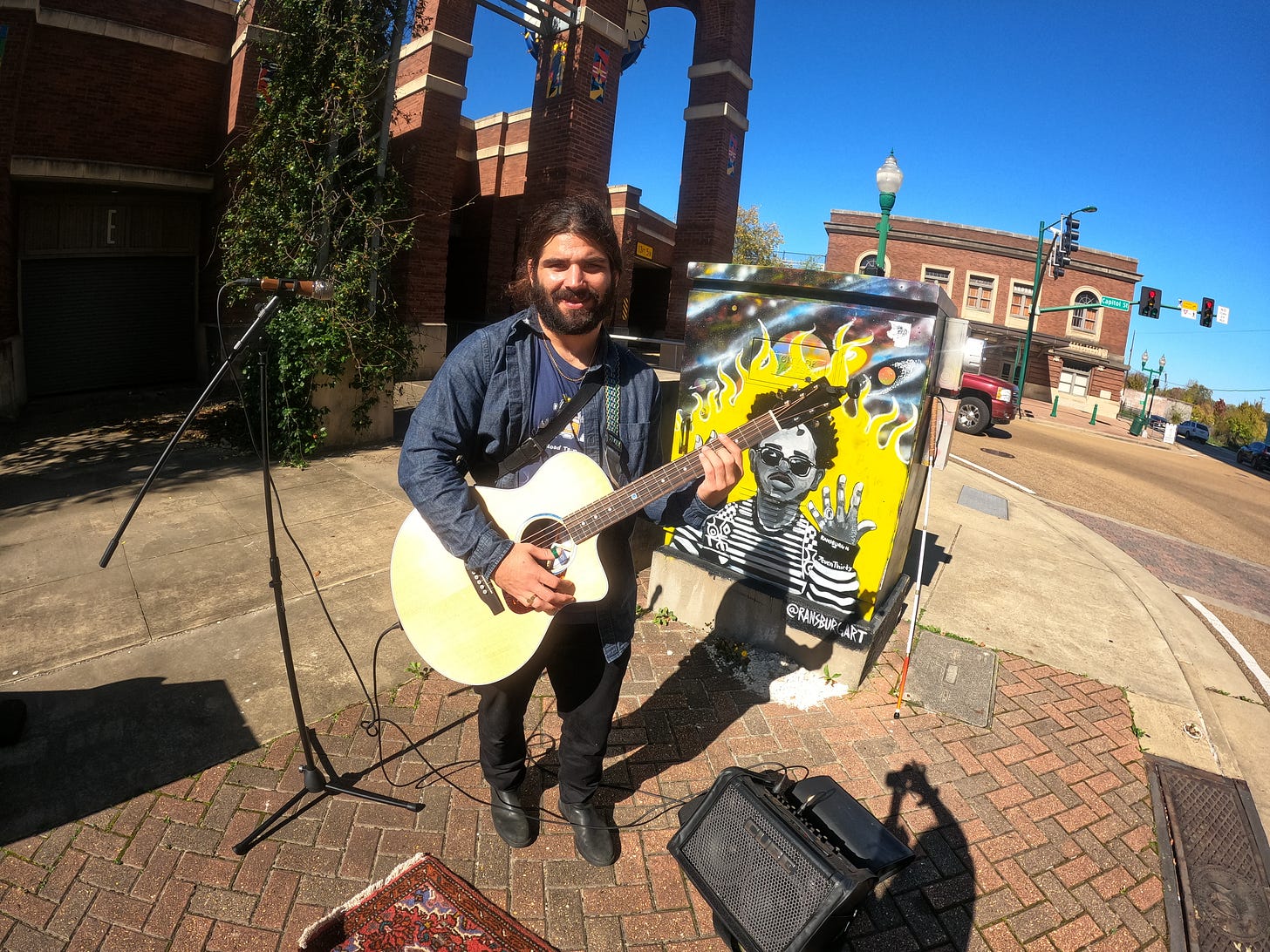 Anthony posted up on the corner getting ready to live stream to Facebook in Jackson, Mississippi a piece of art work is behind him with a guy released from jail in a yellow flame and sunglasses