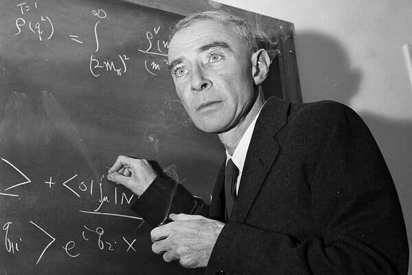 A black-and-white image shows Robert Oppenheimer wearing a dark suit and writes an equation on a chalkboard, his gaze trained on something out of frame.