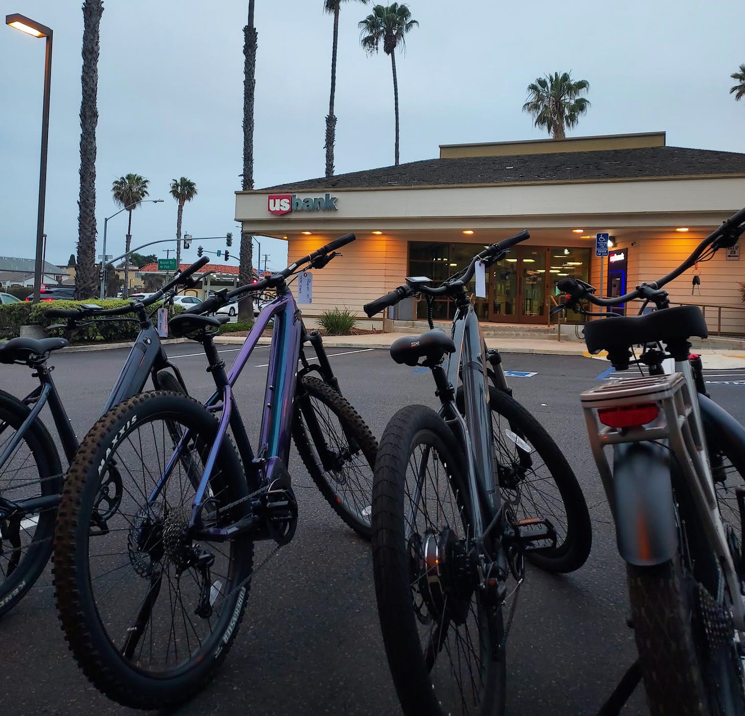 A view of four e-bikes lined up side by side from behind. In the background is a US Bank office set against a dawn sky and palm trees.