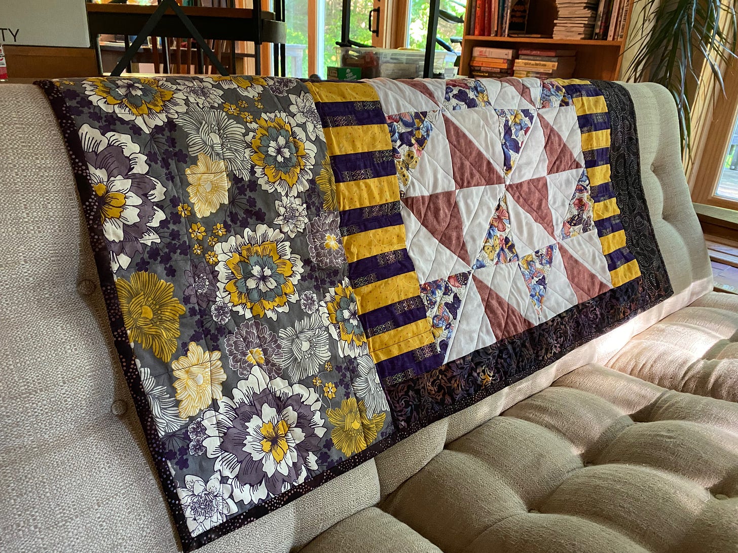 A small rectangular quilt rests on the back of a white sofa. It has contrasting panels of fabrics in shades of purple, blue, white, pink and yellow. There are large flowers, purple and yellow stripes, white and pink triangles, and colorful fabric patterned with butterflies.