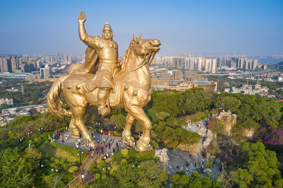 People walk around at the base of an enormous statue of a man riding on a horse.