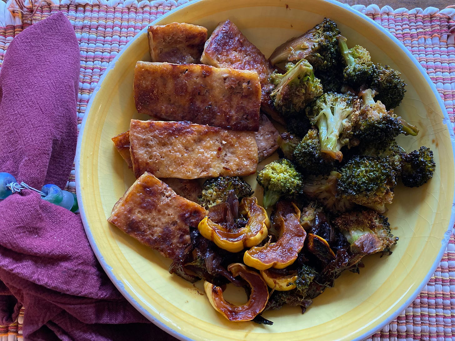 A wide yellow bowl of crispy, browned tofu, slices of roasted delicata squash, and roasted broccoli sits on a checked placemat next to a napkins.