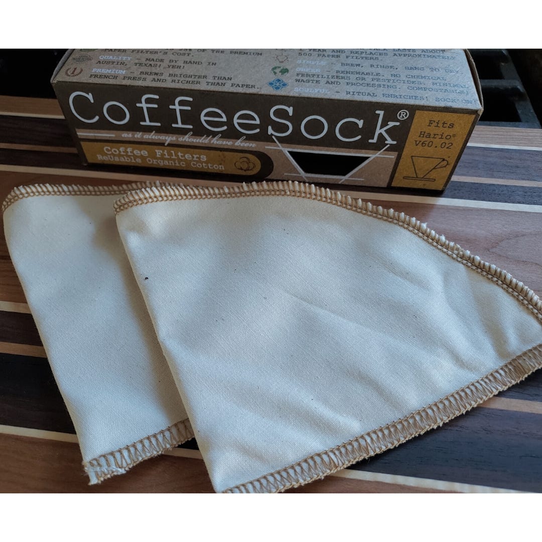 Two triangle shaped cloth coffee filters lying on a wooden cutting board. They are flat and the box they came in with the label "Coffee Sock" is at the top of the image.