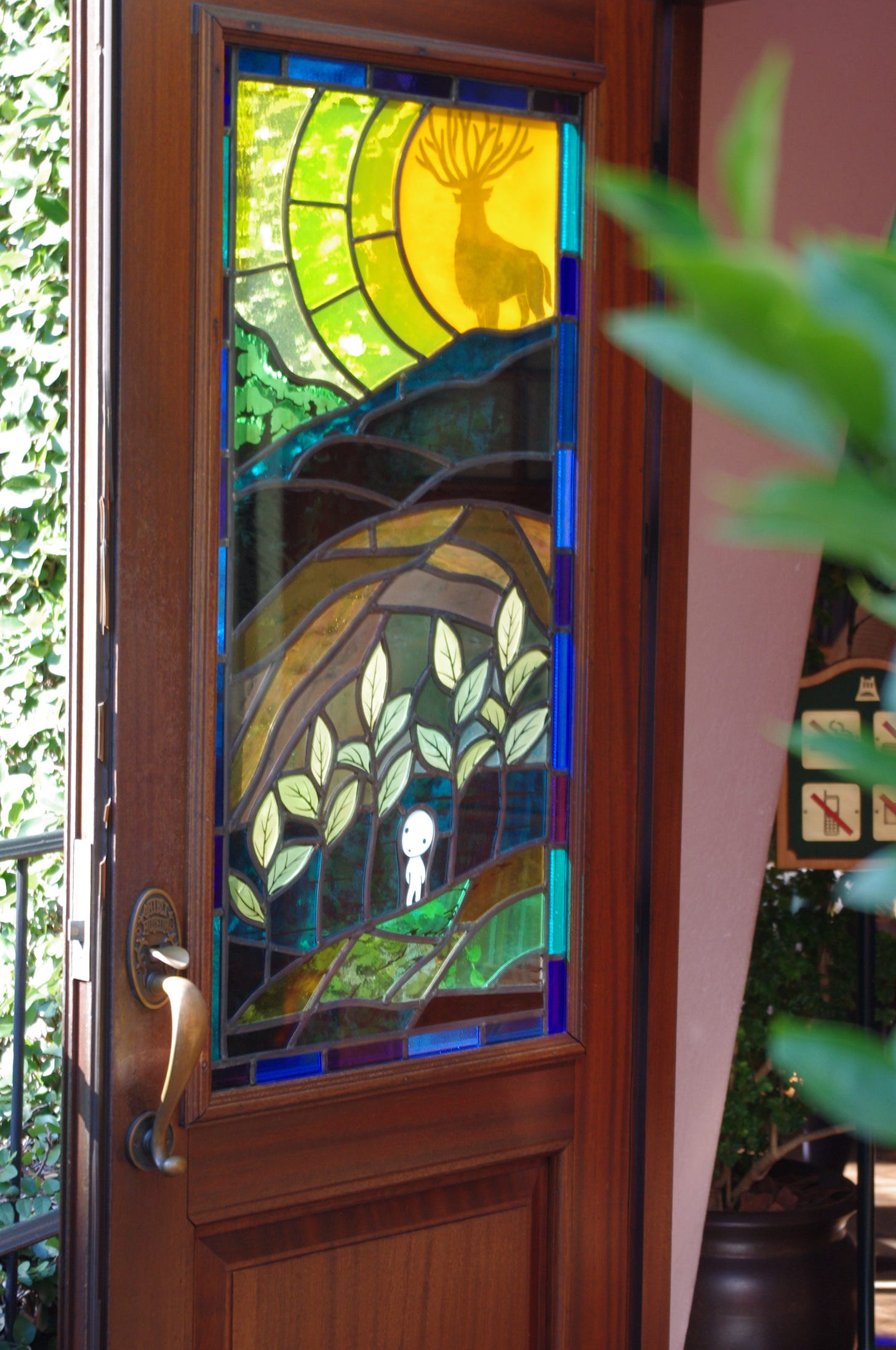 A photograph of a doorway with a stained glass window in it. The subject of the art is the deer from The movie "The Princess Mononoke" (Mononoke-hime) with one of the little click-clack headed spirits at the bottom.
