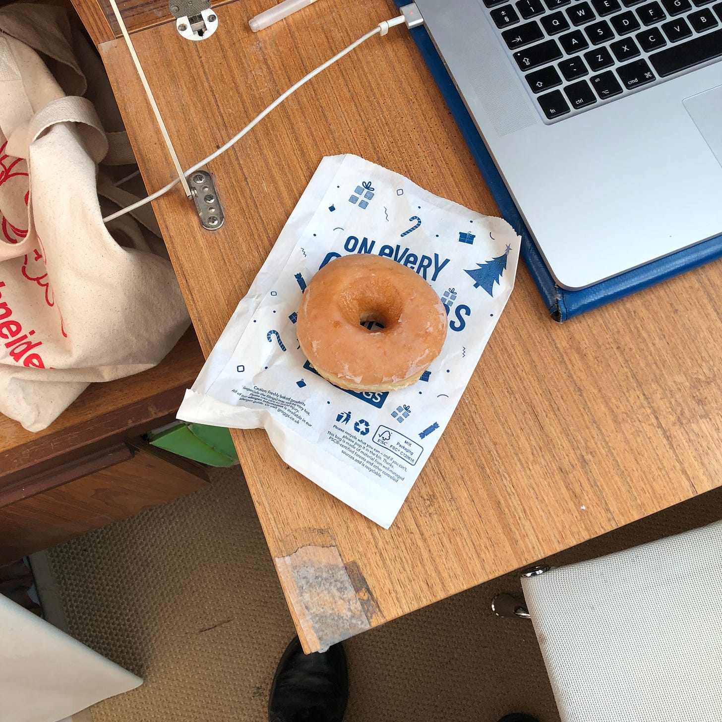 A shot from above of a desk, with a laptop top right. Set out atop a greggs christmas themed paper bag is a classic glazed donut. Cables and a tote bag are around the desk. 