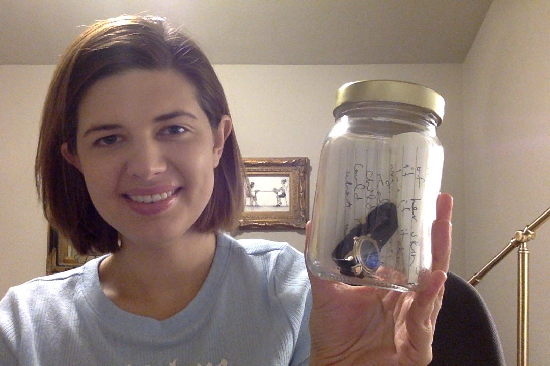 A photo of an ABC volunteer with light skin and shrt, straight brown hair to her neck, wearing a light blue t-shirt and holding a glass jar with some pper and a watch inside. She is sitting in an office with being walls and a painting behind her in a gold frame. 