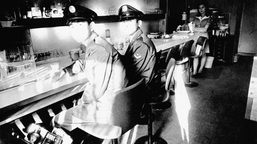 Fotografia em preto e branco de dois oficiais japoneses sentados no balcão de um café como os de filmes americanos. A luz está sobre o rosto deles, mas o contraste é tão alto que não é possível ver seus detalhes. Mais ao fundo, em direção à escuridão, uma garçonete japonesa está encostada no balcão. Seu rosto, nas sombras, já é mais visível: está séria